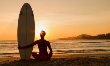 "A girl sitting on the sand with her surfboard, watching the sunset."