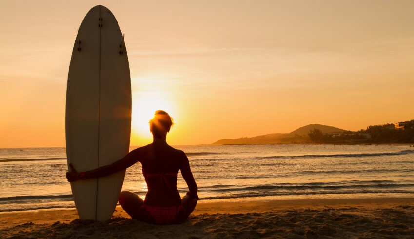 "A girl sitting on the sand with her surfboard, watching the sunset."